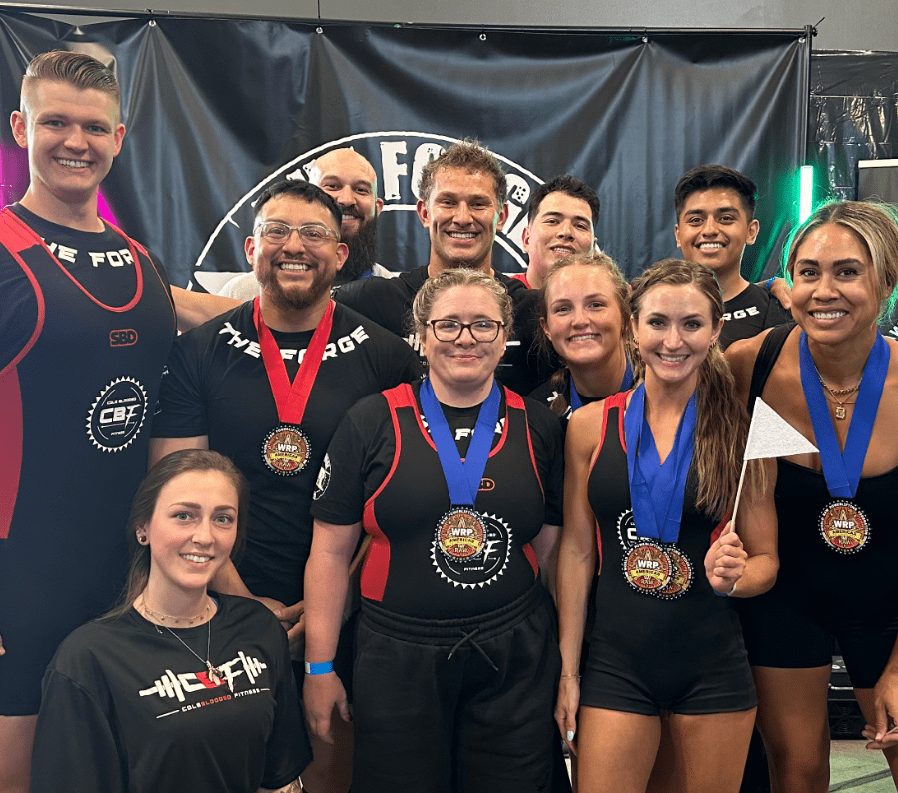 Eleven members of The Forge Gym in team singlets and shirts standing together in front of a Forge banner at a powerlifting meet.