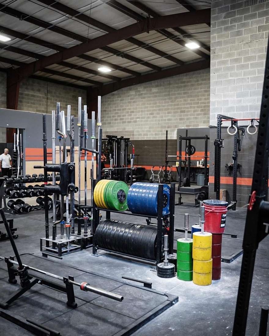 A rack row at The Forge Gym with bar storage, a stack of green, blue and yellow bumper plates, and the orange stripe painted along the block wall.